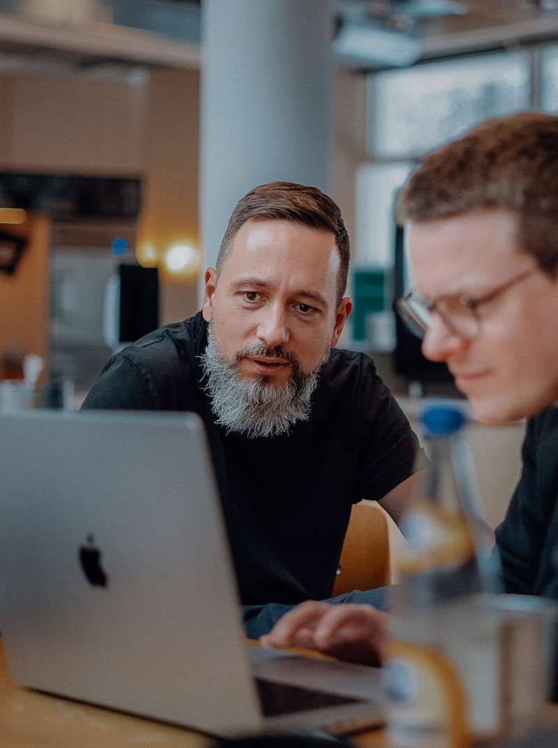 Arne und Markus sitzen gemeinsam im Coworking-Space. Markus arbeitet am MacBook und Arne schaut zu.